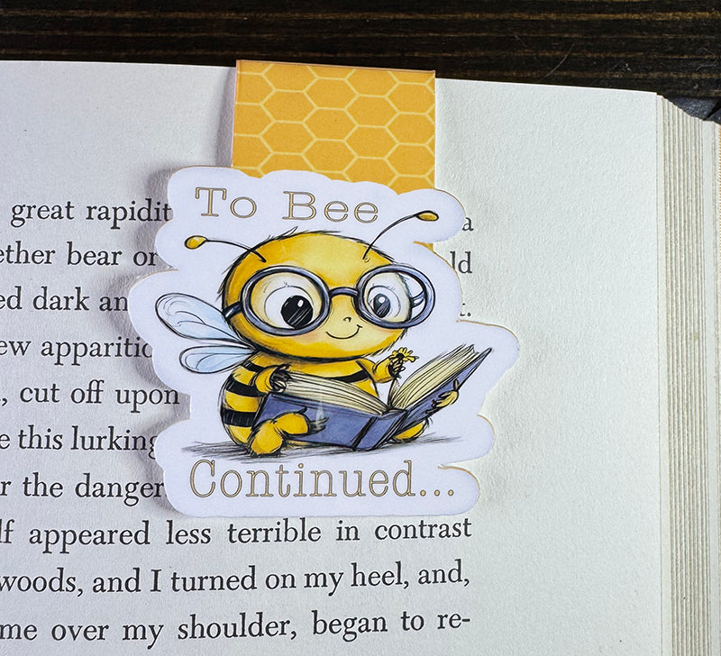 Cute magnetic bookmark with cartoon bee in glasses holding a book and the phrase “To Bee Continued…”