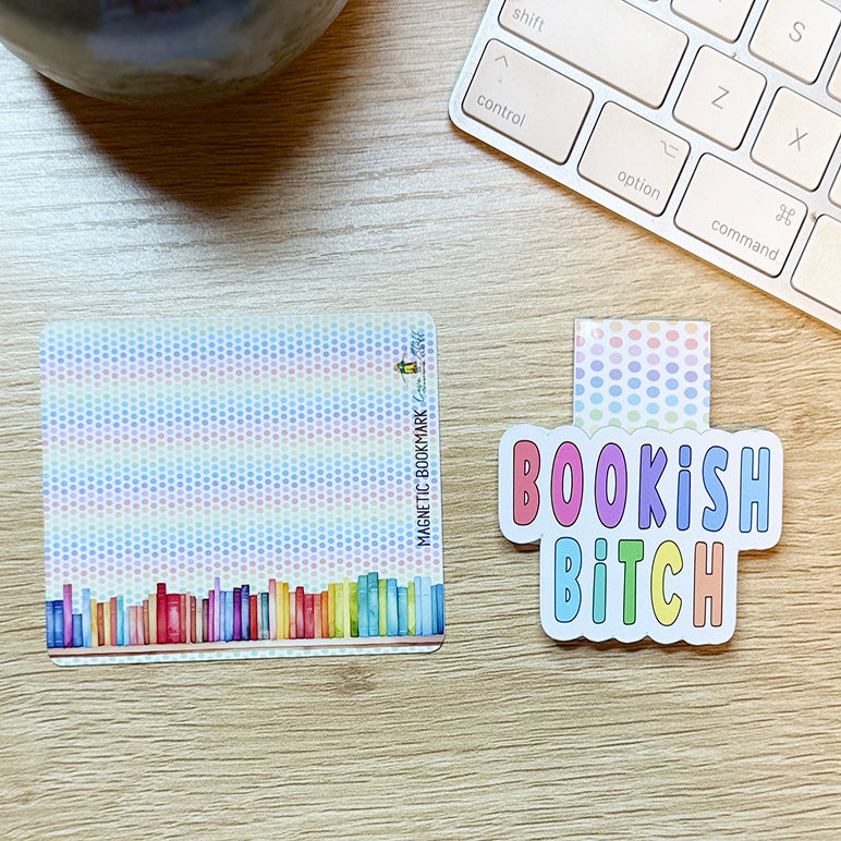 “Bookish Bitch magnetic bookmark with pastel polka-dot tab shown next to its rainbow bookshelf backing card on a light wood surface.”