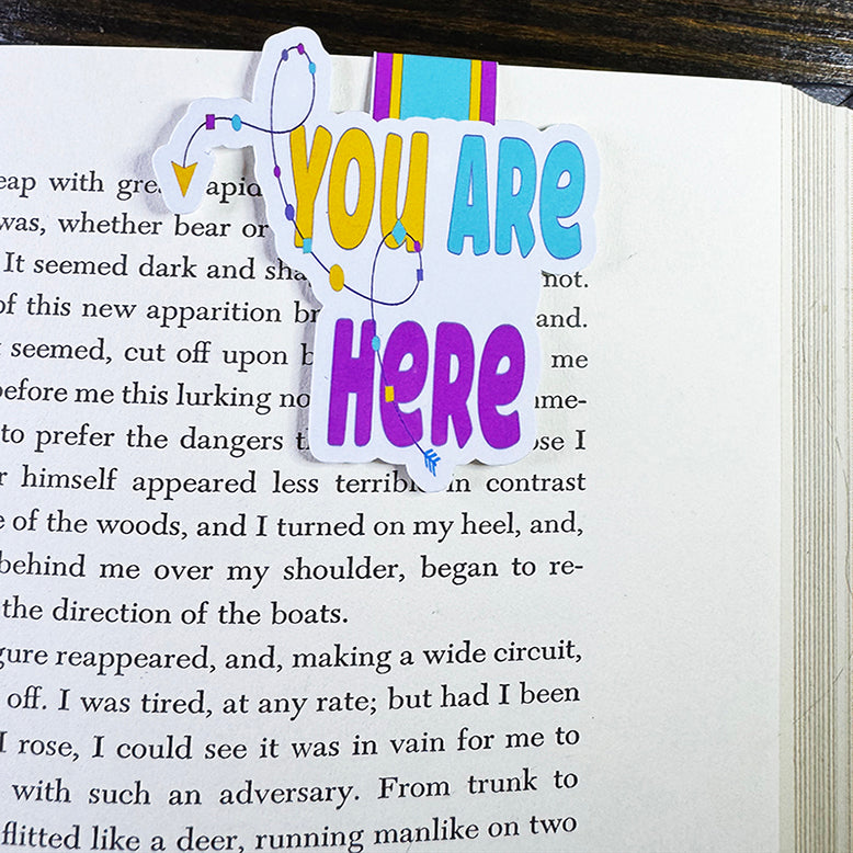 You Are Here magnetic bookmark clipped to the top edge of a book page, marking reading spot with bright and playful design.