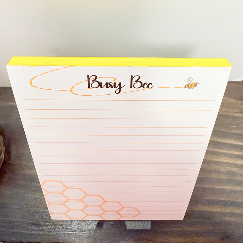 Overhead view of a Busy Bee 5x7 notepad with yellow-edged pages, lined paper, and a cute bee illustration at the top beside the “Busy Bee” title. The notepad has a honeycomb design in the corner and sits on a wooden surface for a warm, home-office aesthetic.