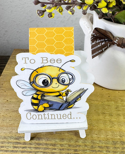 Cute magnetic bookmark with cartoon bee in glasses holding a book and the phrase “To Bee Continued…”