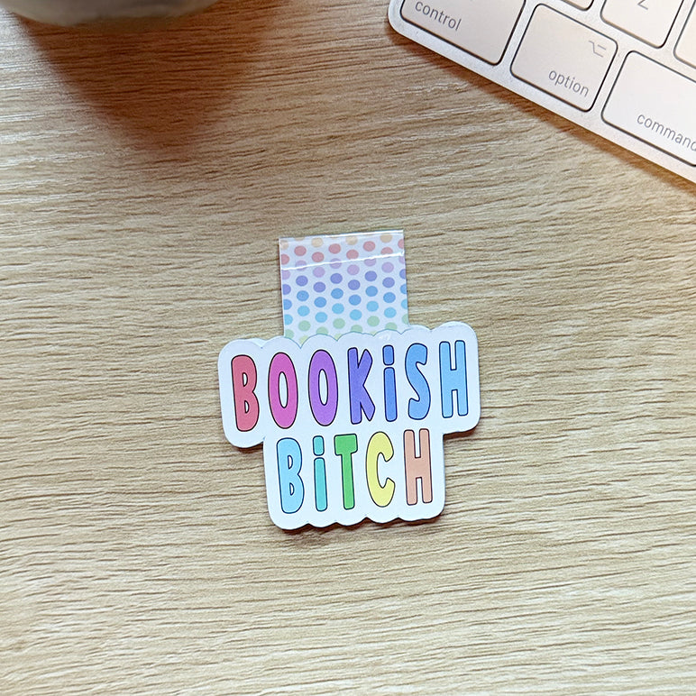 “Colorful magnetic bookmark reading ‘Bookish Bitch’ in bold rainbow letters with pastel polka-dot background, displayed on a wooden desk beside a keyboard.”