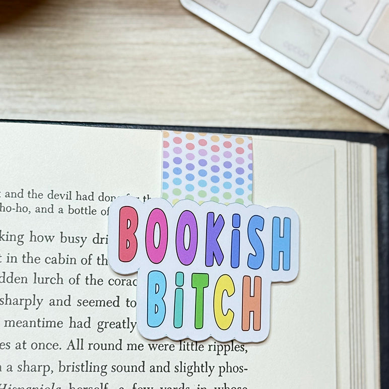 “Bookish Bitch rainbow magnetic bookmark clipped onto an open book page, showing pastel polka-dot tab and colorful bold lettering.”