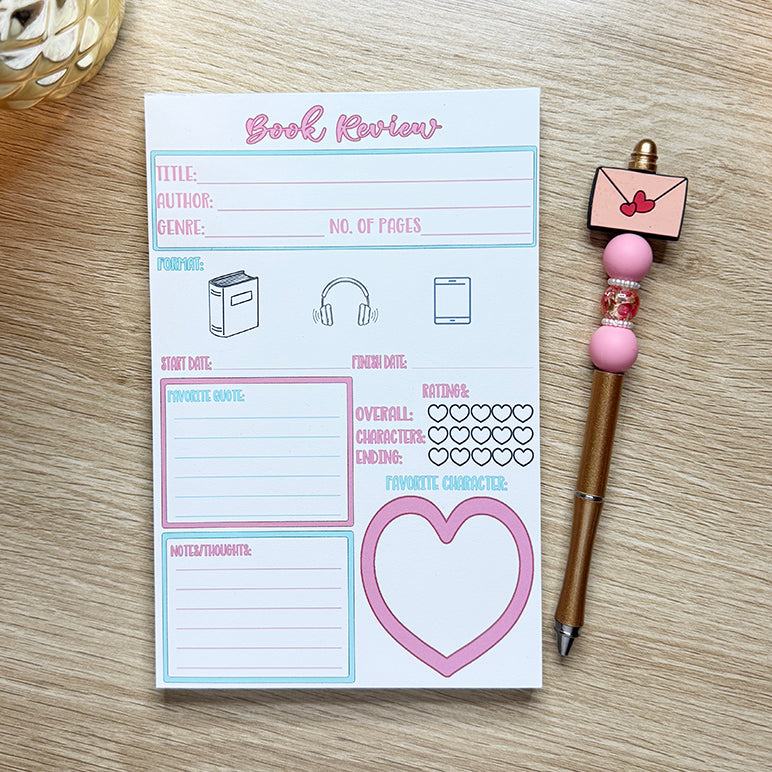 “Close-up of 5x7 book review notepad featuring sections for title, author, favorite quote, and character ratings with heart icons, placed beside a pink pen.”