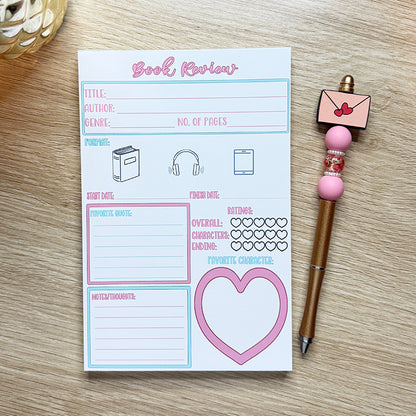 “Close-up of 5x7 book review notepad featuring sections for title, author, favorite quote, and character ratings with heart icons, placed beside a pink pen.”