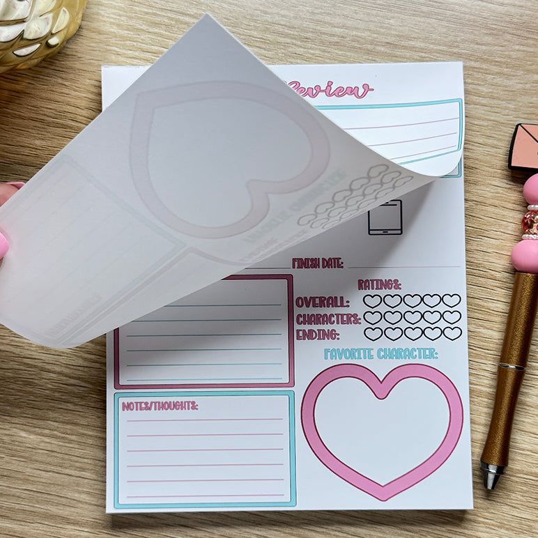 “Book review notepad with pastel pink and blue sections showing tear-off pages and heart-shaped rating design, held open on a wooden desk.”