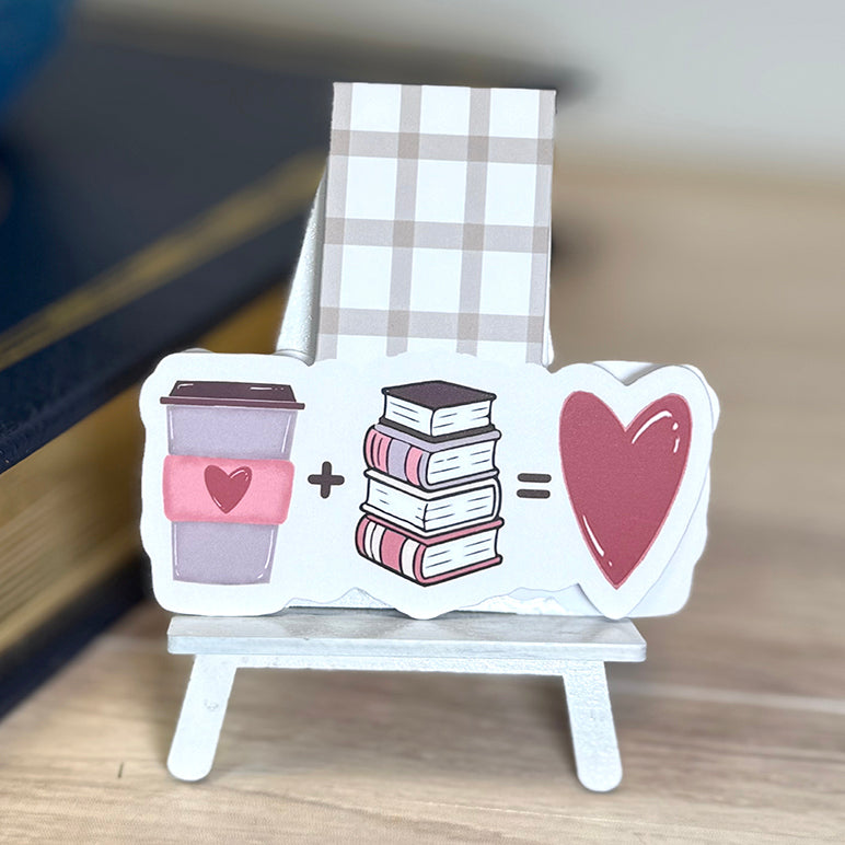 “Close-up of coffee and books magnetic bookmark with illustrated latte, book stack, and heart, attached to neutral plaid magnetic tab.”