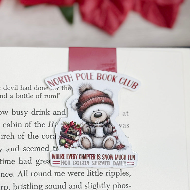 “North Pole Book Club magnetic bookmark clipped on open book showing Christmas bear design and cozy winter quote.”