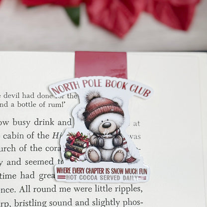 “North Pole Book Club magnetic bookmark clipped on open book showing Christmas bear design and cozy winter quote.”