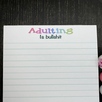 5x7 funny notepad with the phrase “Adulting Is Bullshit” at the top, lined pages, 50 sheets – part of the Notions notepad collection. Close up view of saying.