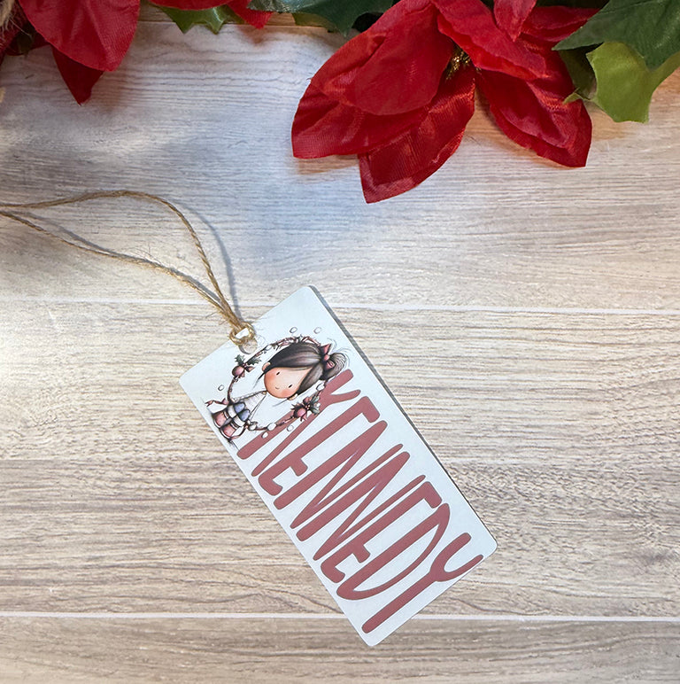 Flat lay of a Christmas bag tag personalized with the name Kennedy and featuring a festive girl surrounded by a holiday wreath.
