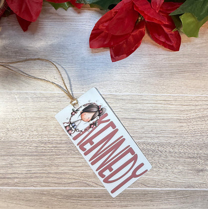 Flat lay of a Christmas bag tag personalized with the name Kennedy and featuring a festive girl surrounded by a holiday wreath.