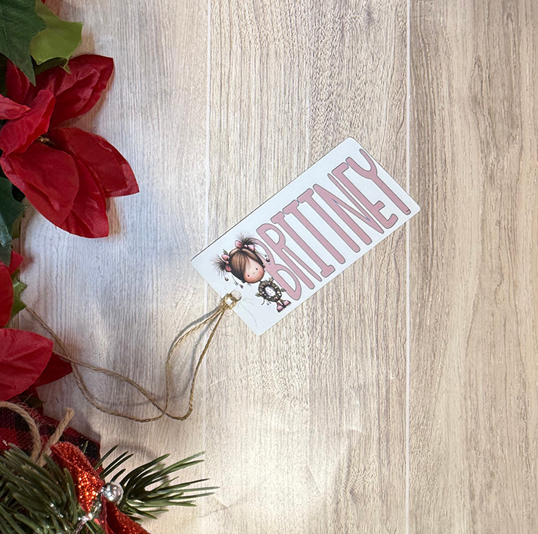 Flat lay of a personalized Christmas tag with the name Brittney and a cute girl character, tied with twine and surrounded by poinsettia leaves.