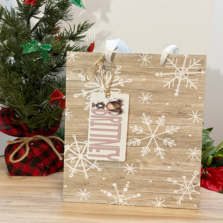 Christmas gift bag with personalized tag reading “Brittney” featuring a cute holiday girl character, tied with twine and displayed next to a small Christmas tree.