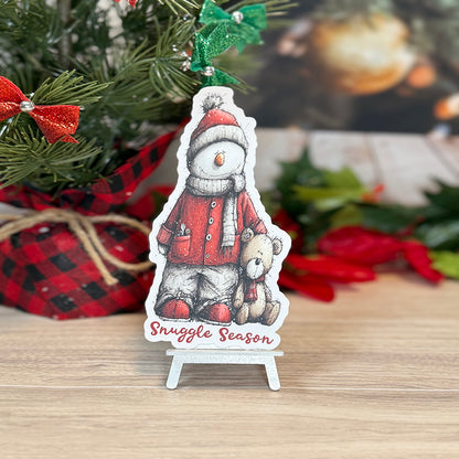 “Classic Snuggle Season bookmark standing on a small display easel, showcasing a winter snowman and teddy bear design with holiday decorations behind it.”