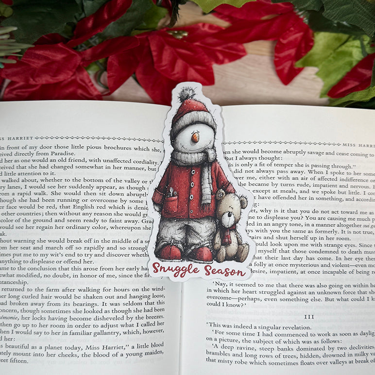 “Classic bookmark featuring a snowman in a red coat and hat holding a teddy bear, sitting between the pages of an open book with holiday décor in the background.”