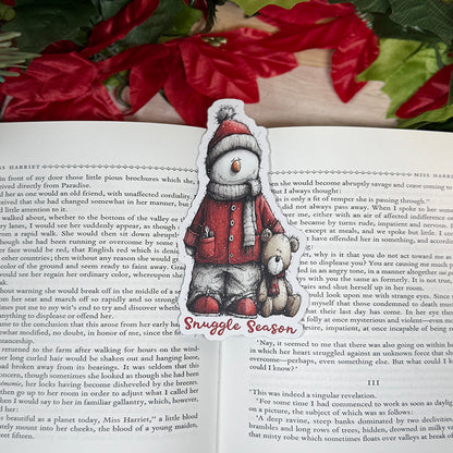“Classic bookmark featuring a snowman in a red coat and hat holding a teddy bear, sitting between the pages of an open book with holiday décor in the background.”