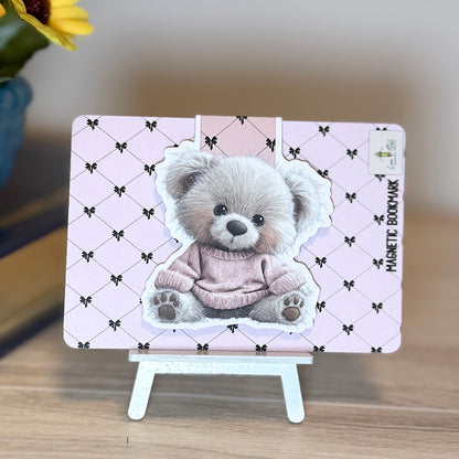 “Teddy bear magnetic bookmark attached to pink backing card with black bow pattern, displayed on small stand.”