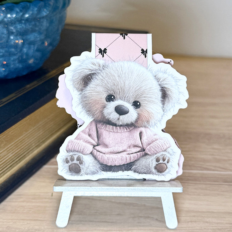 “Magnetic bookmark featuring a fluffy teddy bear in a pink sweater displayed upright on a small stand.”