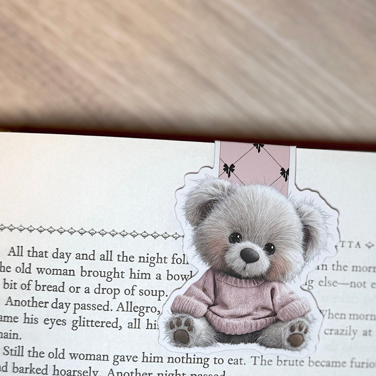 “Pink sweater teddy bear magnetic bookmark clipped to the top of a book page with pink bow-pattern tab visible.”