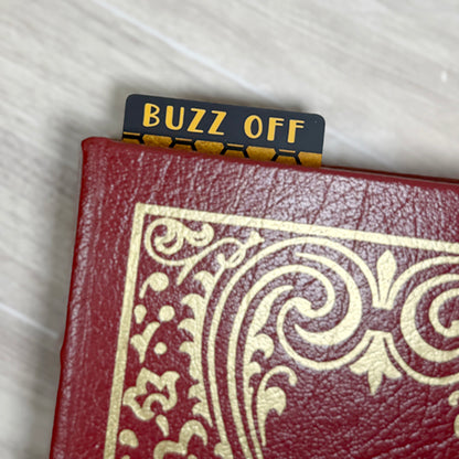 “Buzz Off classic bookmark tab peeking from the top of a closed book with bold yellow text and honeycomb design.”