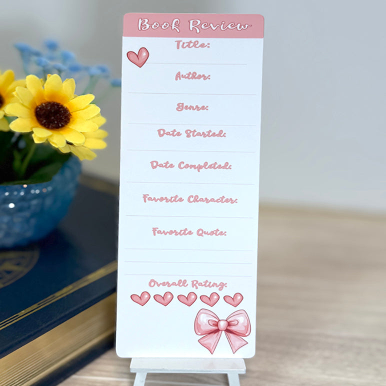 “Book review bookmark standing upright with pink accents, heart icons, and lines for title, author, genre, dates, and rating.”