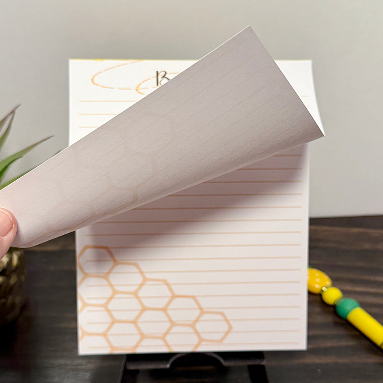 Close-up of a Busy Bee notepad with a honeycomb pattern and lined pages, showing one sheet being lifted to reveal the thickness of the pad. and the tear off sheets. The design features soft golden-yellow tones and a clean minimalist layout, perfect for note-taking or to-do lists.