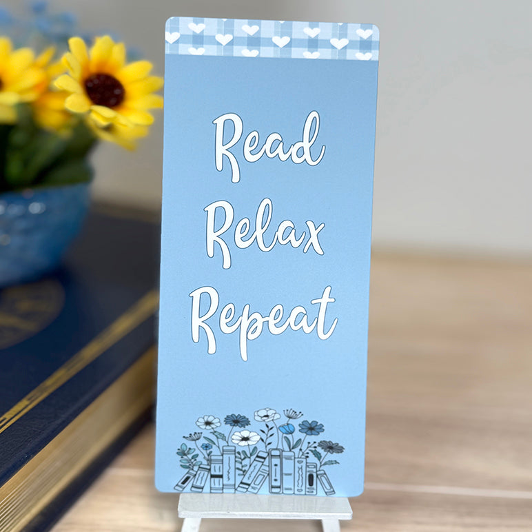 “Blue classic bookmark with the phrase ‘Read Relax Repeat’ and floral book illustration displayed on a small stand.”