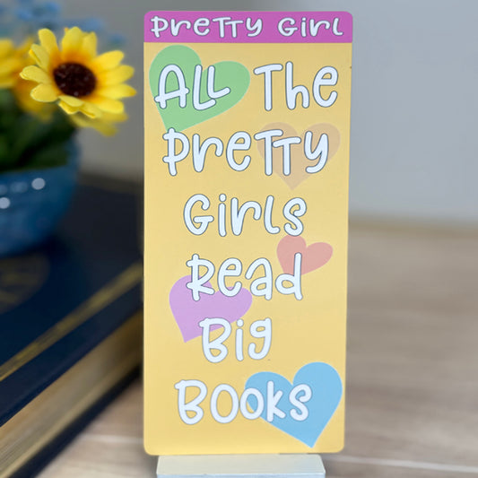 “Standing view of classic bookmark with pastel hearts and the quote ‘All The Pretty Girls Read Big Books’ displayed upright near flowers and books.”