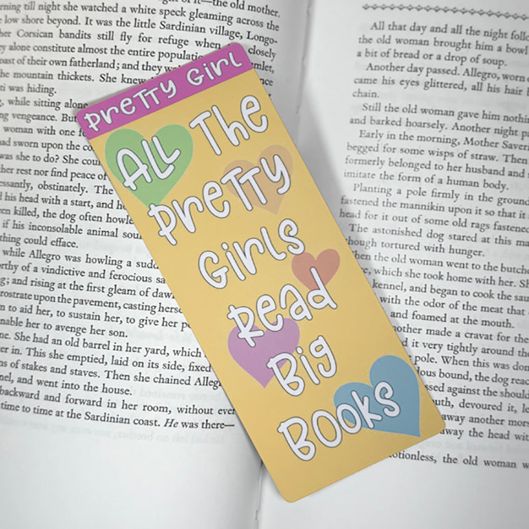 “Classic bookmark with quote ‘All The Pretty Girls Read Big Books’ placed on the pages of an open book with pastel hearts on a yellow background.”