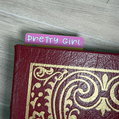 “Top view of ‘Pretty Girl’ bookmark tab peeking out of a closed book with pink and purple lettering.”