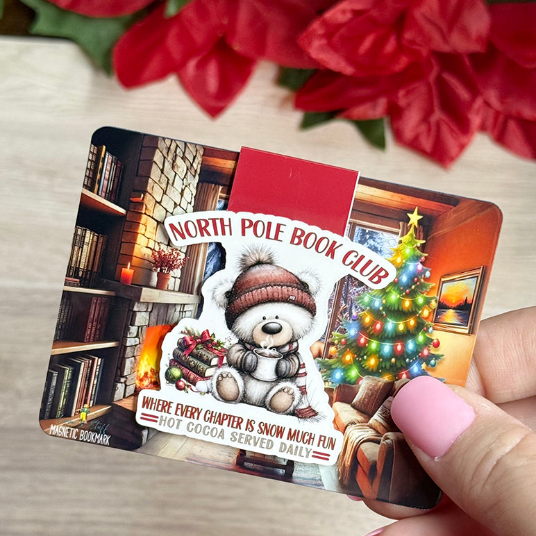 “Close-up of North Pole Book Club magnetic bookmark on Christmas fireplace backing card with holiday tree.”