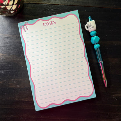 Flat lay of the Pink Bow Notes Notepad with a teal and pink border and a “Girl Boss” pen beside it, perfect for everyday note-taking.