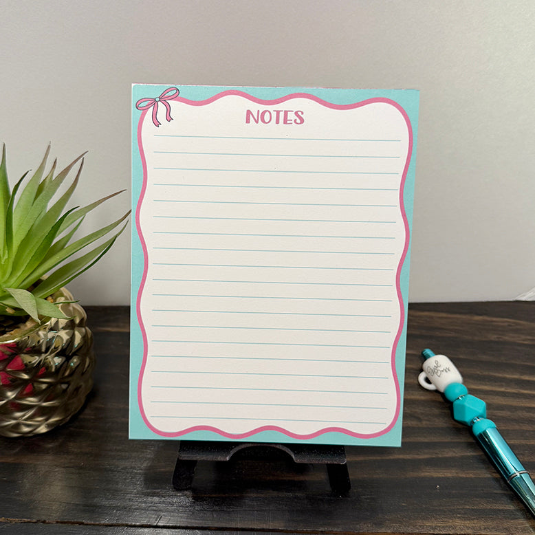 Front view of Pink Bow Notes Notepad with teal border, pink scalloped edges, and lined pages, displayed on a wooden surface beside a teal beaded “Girl Boss” pen.