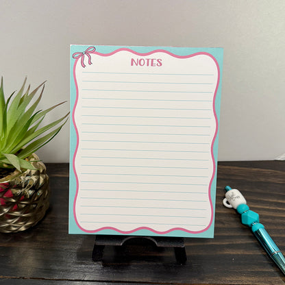 Front view of Pink Bow Notes Notepad with teal border, pink scalloped edges, and lined pages, displayed on a wooden surface beside a teal beaded “Girl Boss” pen.